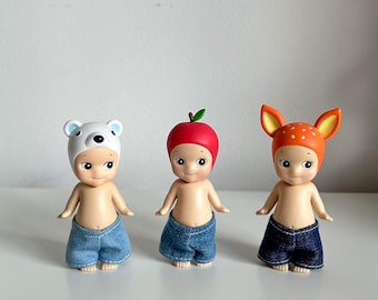 May include: Three small plastic dolls with different hats and blue denim shorts. The dolls are wearing a white polar bear hat, a red apple hat, and an orange deer hat.
