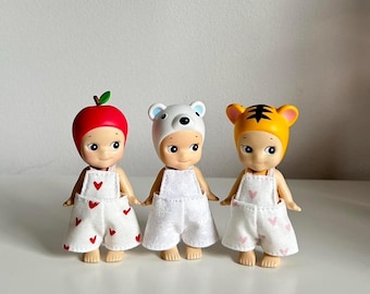 May include: Three small dolls wearing overalls and different hats. The doll on the left is wearing a red apple hat, the middle doll is wearing a white bear hat, and the doll on the right is wearing a yellow tiger hat.
