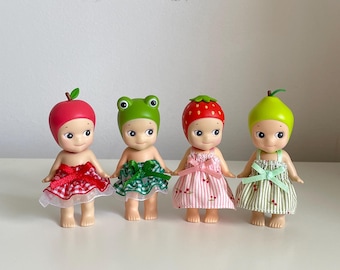 May include: Four small dolls dressed in fruit-themed outfits. The dolls are wearing red and white checkered, green and white checkered, pink and white striped, and green and white striped dresses. The dolls have different fruit-shaped hats, including an apple, a frog, a strawberry, and a pear.