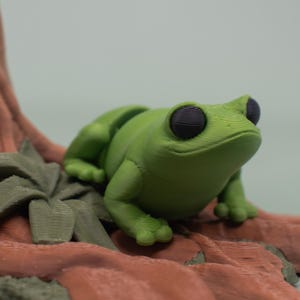 May include: A green frog figurine with black eyes sits on a brown, textured base. The frog is posed with its legs bent and its head tilted up.