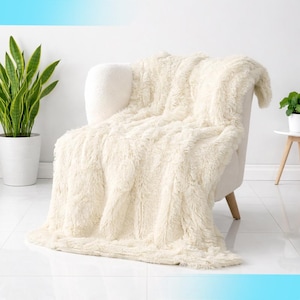 May include: A cream-colored, fluffy faux fur throw blanket draped over a white armchair. The blanket has a long, shaggy texture. A potted snake plant and a small side table with a plant are visible in the background.
