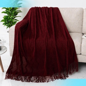 May include: A burgundy-colored throw blanket with a textured pattern and fringe detail draped over a beige sofa. The blanket's intricate design and rich color create a cozy and inviting aesthetic. The throw blanket is a decorative home accessory.