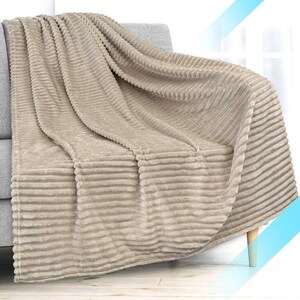 May include: A beige, ribbed throw blanket draped over a gray sofa. The blanket has a soft, plush texture and a horizontal striped pattern. The sofa has light blue legs.