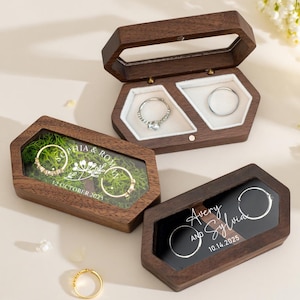 Op de afbeelding: Drie zeshoekige houten ringendoosjes met doorzichtige deksels. Eén doosje is open en toont twee ringen. De andere twee doosjes zijn gesloten, met de tekst "Sophia & Rom" en "Avery and Sylvia" met data.