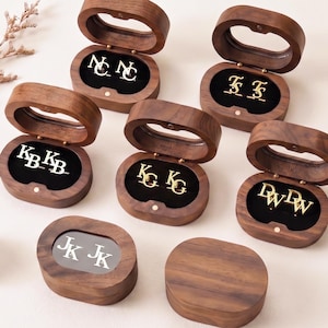 May include: Six oval dark wood boxes with hinged lids, five open to reveal gold or silver metal cufflinks with engraved initials including NC, TS, KB, KG, and DW. One closed box shows the initials JK on a silver plate. Pinecones are placed nearby.