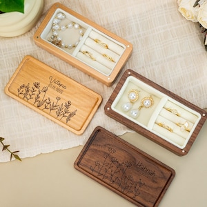 May include: Wooden jewelry boxes with personalized floral engravings and names. One box is light wood, another is dark wood. The boxes contain jewelry such as rings, earrings, and a pearl necklace. The text "Victoria 07.19.2009" and "Hannah 03.19.2023" are engraved on the boxes.