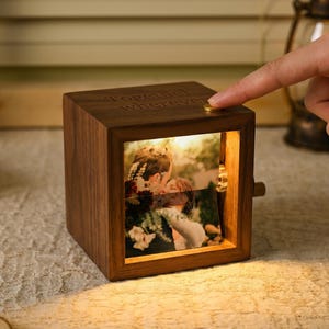May include: A wooden photo cube with a warm glow, displaying a couple's photo. The cube has the words "Together Wherever" engraved on top. A finger presses a gold button on the cube. The cube is illuminated from within.