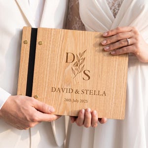 Peut inclure: Livre d'or de mariage en bois clair, gravé avec les noms "David & Stella" et la date du "26 juillet 2025". Le livre a une tranche noire et est tenu par deux personnes, l'une en costume blanc et l'autre en robe blanche.