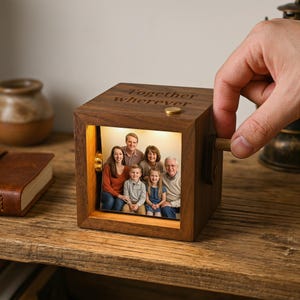 May include: A wooden photo cube displaying a family portrait, lit from within. The cube is engraved with the words "Together wherever". A hand turns a wooden crank. The cube rests on a wooden surface, with a small book and vase nearby.
