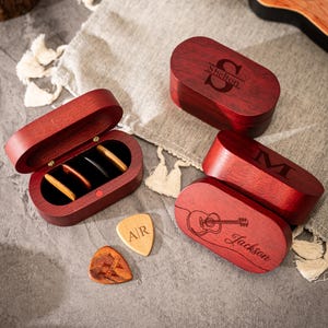 Op de afbeelding: Een verzameling gepersonaliseerde houten gitaar plectrumdozen en plectrums. De dozen zijn dieprood van kleur, met gegraveerde namen en initialen. Eén doos is open en onthult gitaar plectrums. Een ukelele staat op de achtergrond.