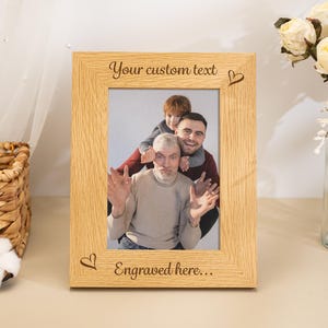 Puede incluir: Un marco de fotos rectangular de madera clara con un patrón de veta visible. El borde superior está grabado con el texto "Your custom text" y un corazón, mientras que el borde inferior presenta el texto "Engraved here..." y un corazón.