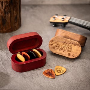 Puede incluir: Una caja roja de púas de guitarra de madera con varias púas dentro. Una caja de madera con notas musicales y las iniciales "W.S" está al lado de la caja de púas. Dos púas de guitarra están sobre la superficie.