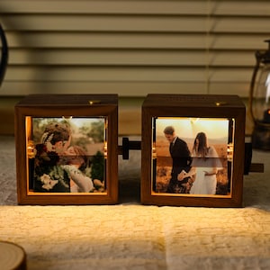 May include: Two wooden photo cubes displaying illuminated images of a couple. The cubes are linked and have a crank. Warm light emanates from within, highlighting the photos. A lantern and headphones are in the background.