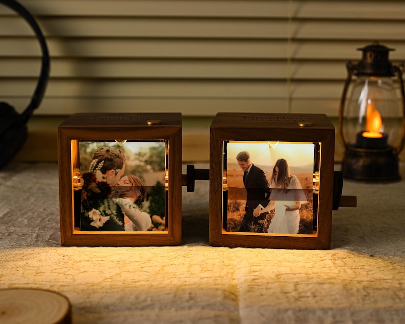 May include: Two wooden photo cubes displaying illuminated images of a couple. The cubes are linked and have a crank. Warm light emanates from within, highlighting the photos. A lantern and headphones are in the background.