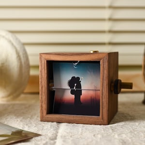 May include: A wooden photo cube with a rotating image of a couple silhouetted against a sunset. The cube has a dark brown finish and a brass knob. A decorative lantern and a white object are in the background.