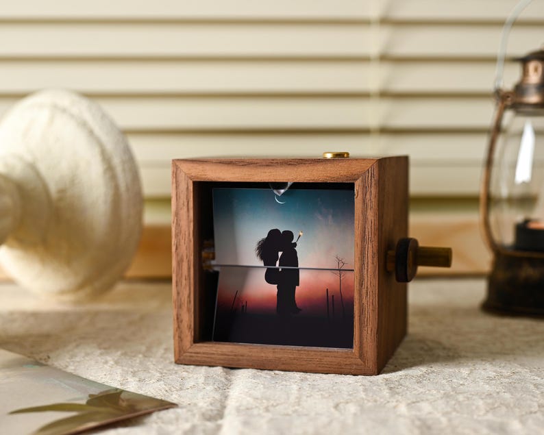 May include: A wooden photo cube with a rotating image of a couple silhouetted against a sunset. The cube has a dark brown finish and a brass knob. A decorative lantern and a white object are in the background.