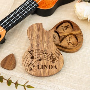 Puede incluir: Un soporte para púas de guitarra de madera con forma de guitarra, con un diseño de notas musicales y el nombre "Linda" grabado en la parte delantera. El soporte tiene tres ranuras para púas de guitarra, una de las cuales está grabada con el nombre "Kevin Stewart" y una cabeza de ciervo.