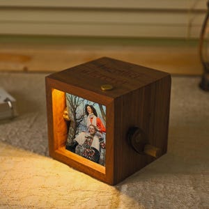 May include: A wooden music box in a cube shape, with a photo of two people visible. The box has a crank on the side and the words "together forever" engraved on top. A small lamp is in the background.