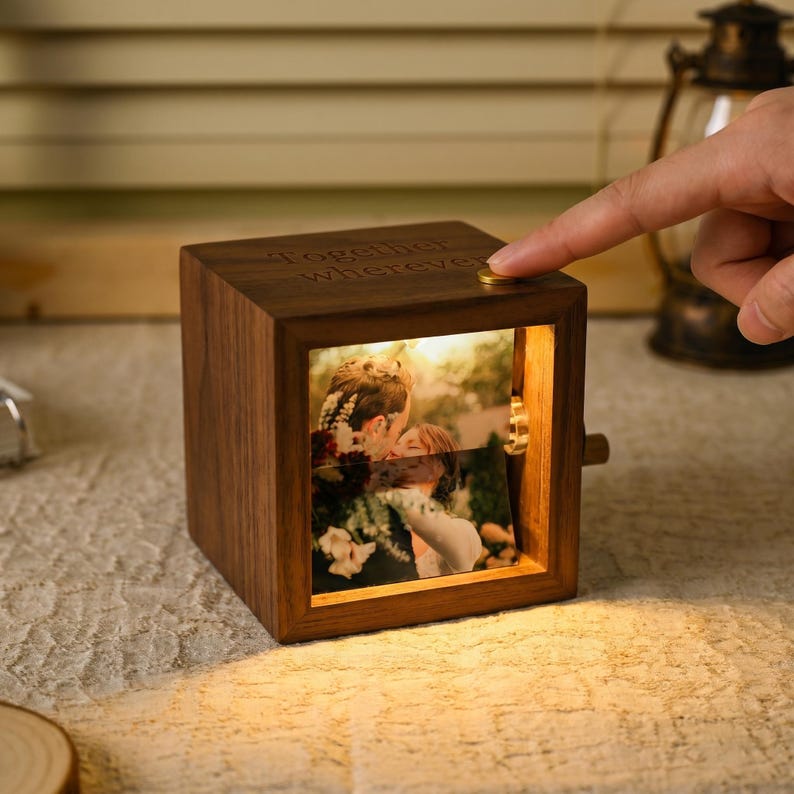 May include: A wooden photo cube with a warm glow, displaying a couple's photo. The cube has the words "Together Wherever" engraved on top. A finger presses a gold button on the cube. The cube is illuminated from within.