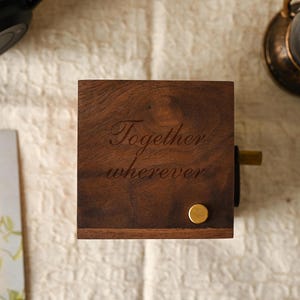 May include: A dark brown wooden music box, engraved with the words "Together wherever". It features a gold-coloured winding knob and a small, round gold-coloured stud. The box sits on a textured, off-white surface.