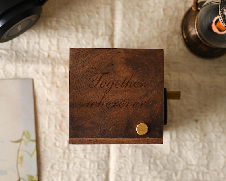May include: A dark brown wooden music box, engraved with the words "Together wherever". It features a gold-coloured winding knob and a small, round gold-coloured stud. The box sits on a textured, off-white surface.