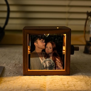 May include: A wooden photo cube displaying a rotating image of a couple, lit from within. The cube emits a warm glow, and a small disco ball is visible in the image. A lantern and headphones are in the background.