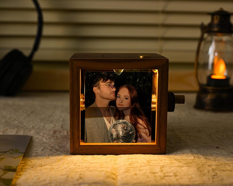 May include: A wooden photo cube displaying a rotating image of a couple, lit from within. The cube emits a warm glow, and a small disco ball is visible in the image. A lantern and headphones are in the background.