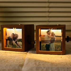 May include: Two wooden photo cubes with illuminated pictures of couples. The cubes have a warm glow, and a small knob on the side. The photos are displayed horizontally, and the wood has a rich, dark tone. The cubes are lit.