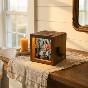 May include: A wooden photo cube displaying a picture of two people, lit from inside. The cube is engraved with the words "Together Wherever". A lit candle sits on a small plate to the left. The cube rests on a wooden table, with a decorative runner.
