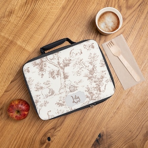 Peut inclure: Un sac à déjeuner blanc avec un imprimé Winnie l'ourson brun. Le sac a une fermeture éclair noire et une poignée noire. Le sac a une étiquette de nom personnalisée qui dit "Lily".