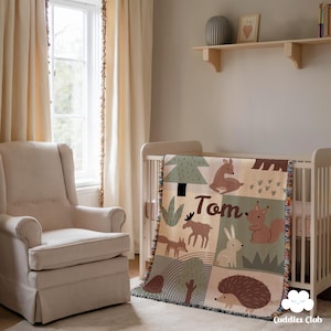 May include: A beige and green baby blanket with woodland animal illustrations, including a deer, squirrel, and hedgehog. The blanket is draped over a white crib and features the name "Tom."