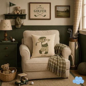 Può includere: Una poltrona beige presenta un cuscino quadrato con un orso ricamato che indossa un completo da golf e tiene in mano una mazza, con il nome Carter sotto. Una coperta a quadri verde e beige è drappeggiata sul bracciolo.