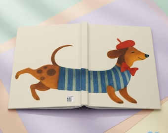 Puede incluir: Diario abierto con una ilustración caprichosa de un perro salchicha. El perro lleva una camisa a rayas azules y blancas, una pajarita roja y una boina roja. El diario está abierto, revelando el diseño ilustrado.