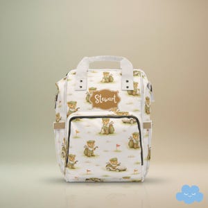 Könnte beinhalten: Weiße Wickeltasche mit einem sich wiederholenden Muster von Golf spielenden Teddybären. Die Tasche hat eine Vordertasche, Seitentaschen und einen oberen Griff. Der Name "Stewart" ist auf einem braunen Etikett aufgedruckt.
