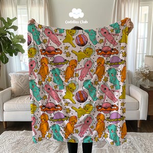 May include: A white fleece blanket with a repeating pattern of colorful cartoon aliens, planets, and stars. The text "Cuddles Club" is printed in the top center of the blanket.