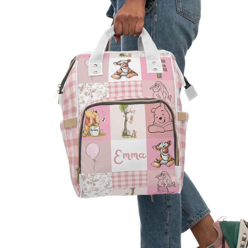May include: Pink and white checkered diaper bag with Winnie the Pooh and Tigger characters. The bag has a large pocket on the front with the name "Emma" printed on it.