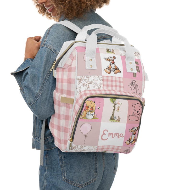 May include: Pink and white diaper bag with a patchwork design featuring Winnie the Pooh characters. The bag has a large pocket on the front with the name "Emma" printed on it.