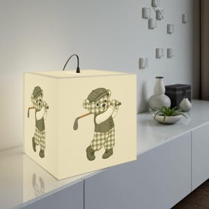 Puede incluir: Una lámpara de mesa cuadrada con una pantalla de color crema que presenta una ilustración repetida de un oso de peluche con gorra, chaleco y pantalones de cuadros verdes mientras sostiene un palo de golf. La lámpara tiene una pequeña asa negra.