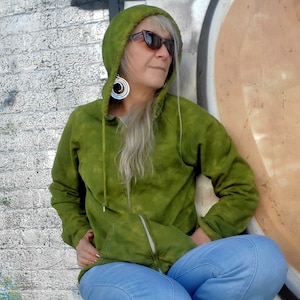 May include: A woman wearing a green tie-dye hoodie with a zipper and a pair of blue jeans. She is wearing sunglasses and large silver hoop earrings.