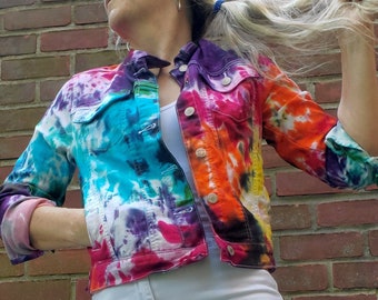 Könnte beinhalten: Eine Jeansjacke im Batik-Look in leuchtenden Farben wie Blau, Lila, Rosa, Orange und Gelb. Die Jacke hat ein klassisches Design mit Knopfleiste, Kragen und Taschen. Kombiniert mit weißen, zerrissenen Jeans.