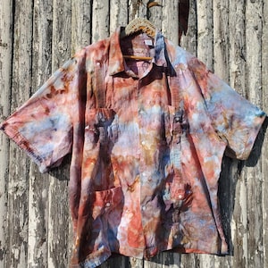 Puede incluir: Una camisa de manga corta con botones y un estampado tie-dye en tonos azules, rojos y marrones. La camisa tiene dos bolsillos en el pecho y un cuello.
