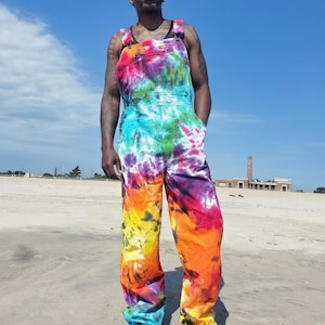 May include: Full-length overalls featuring a vibrant tie-dye pattern in shades of red, orange, yellow, green, blue, and purple. The garment includes a bib pocket with a visible brand logo, adjustable shoulder straps, and side pockets.