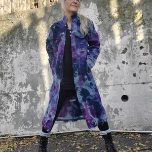 Puede incluir: Una chaqueta vaquera larga con un patrón tie-dye en tonos de morado y azul. La chaqueta tiene un aspecto desgastado con bordes deshilachados y se lleva sobre una camisa negra. La persona que lleva la chaqueta está de pie frente a una pared de hormigón con una valla de madera en el fondo.