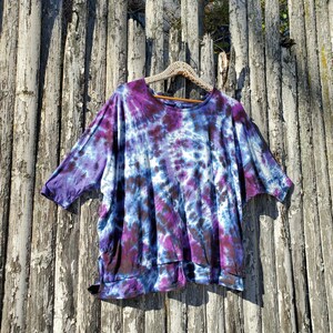 May include: A purple, blue, and black tie-dye shirt with a relaxed fit and short sleeves. The shirt is hanging on a wooden hanger against a weathered wooden fence.