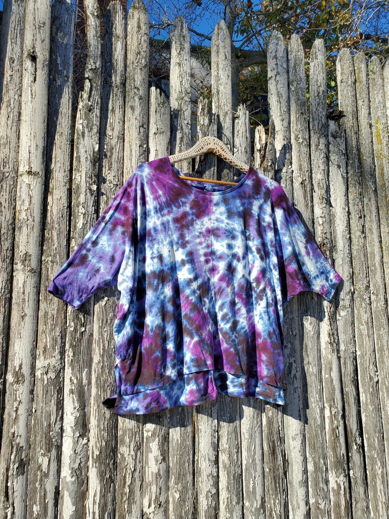 May include: A purple, blue, and black tie-dye shirt with a relaxed fit and short sleeves. The shirt is hanging on a wooden hanger against a weathered wooden fence.