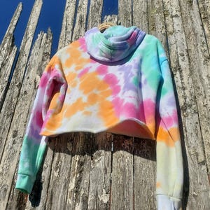 May include: A cropped tie-dye hoodie in a vibrant mix of colors, including pink, orange, purple, and teal. The hoodie features a drawstring hood and long sleeves with white cuffs. The garment is hanging on a wooden fence against a blue sky.