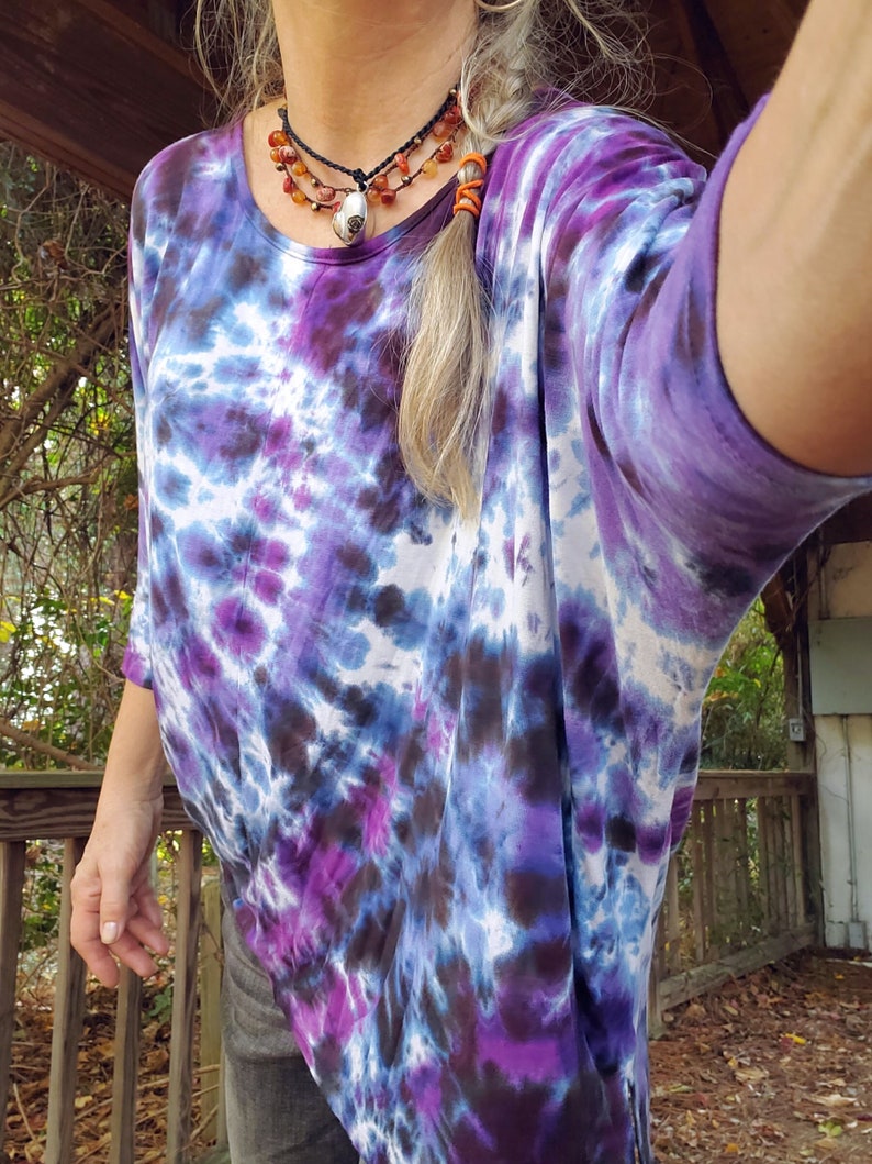 May include: A purple, blue, and white tie-dye shirt with a long, flowing design. The shirt has a scoop neck and three-quarter sleeves. The model is wearing a layered necklace with a silver pendant.