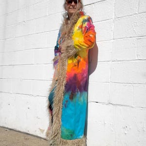 Peut inclure: Un long manteau tie-dye avec un mélange de couleurs vives, dont le jaune, l'orange, le bleu et le violet. Le manteau est orné d'une bordure en fausse fourrure sur les bords et le col. C'est un vêtement unique.