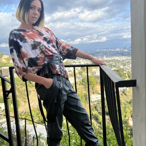 May include: A person wearing a tie-dye shirt with a black belt and black leather pants with suspenders. The person is standing on a balcony overlooking a city.