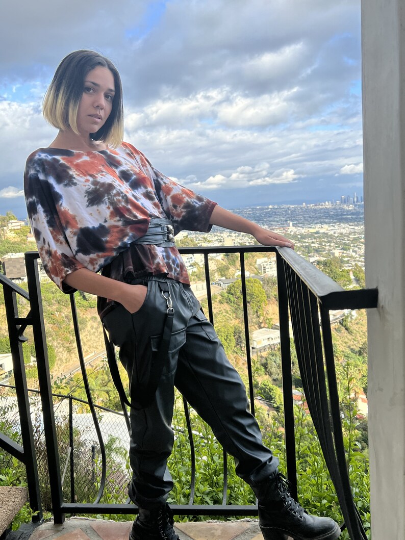May include: A person wearing a tie-dye shirt with a black belt and black leather pants with suspenders. The person is standing on a balcony overlooking a city.